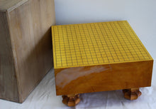Load image into Gallery viewer, A wooden Go board with a yellow grid and black markings, supported by four carved wooden feet, shown next to its matching wooden storage box on a white fabric surface.